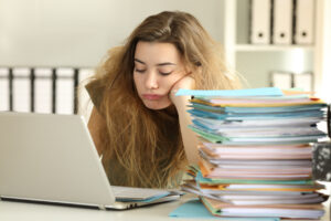 mulher ignorando suas tarefas, dormindo na frente do computador com uma pilha de papel ao seu lado